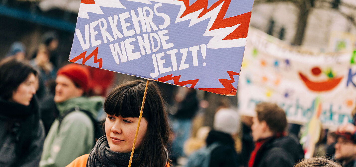 Dossier Junge Menschen Verkehrswende Jetzt! Junge Frau auf einer Demo hält ein Schild in die Luft, auf dem steht "Verkehrswende Jetzt!"